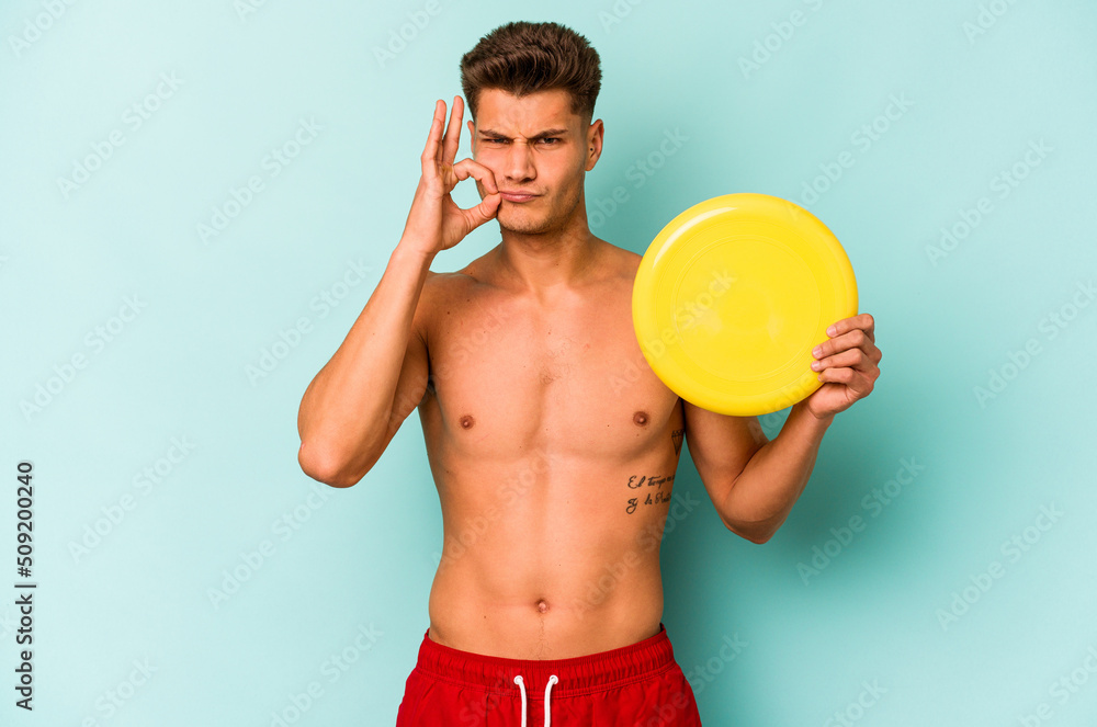 Young caucasian man playing with frisky isolated on blue background with fingers on lips keeping a secret.