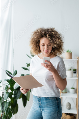 curly woman holding folder with documents while using cellphone at home.