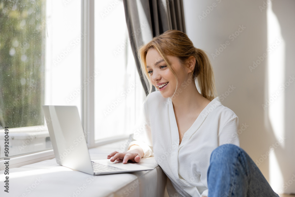 Positive freelancer using laptop on windowsill in living room. Positive freelancer using laptop on windowsill in living room.