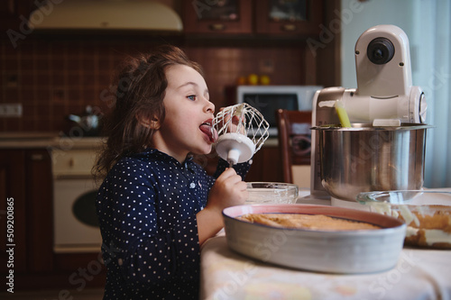 Cute little girl making funny faces while licking whisk with leftovers of whipped cheese cream in steel bowl at home kitchen, standing near a plate with freshly made Tiramisu cake