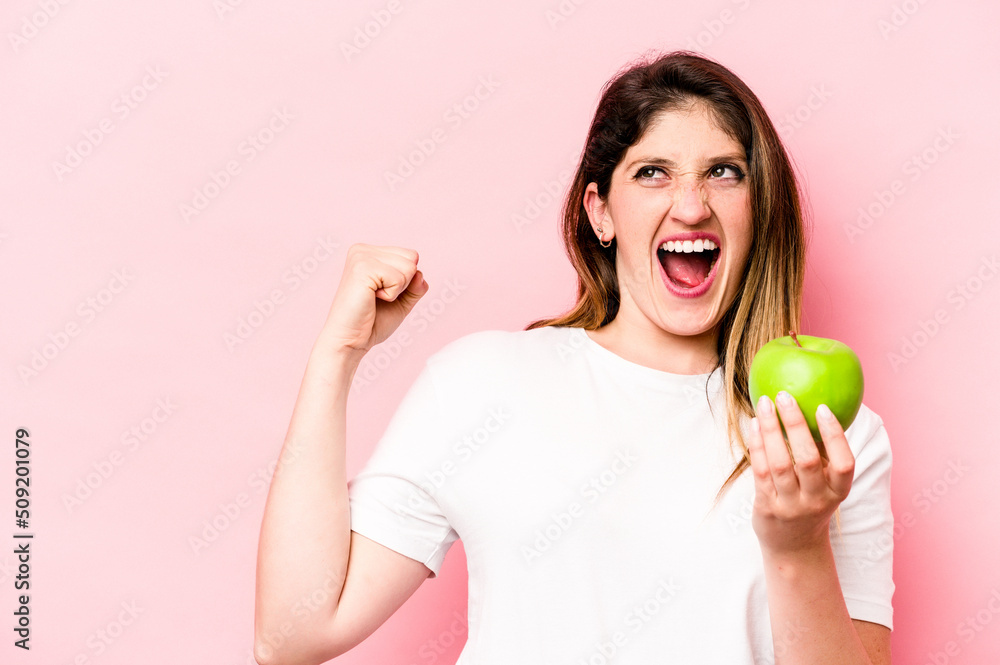 Young caucasian woman holding an apple isolated on pink background raising fist after a victory, winner concept. Young caucasian woman holding an apple isolated on pink background raising fist after a victory, winner concept.