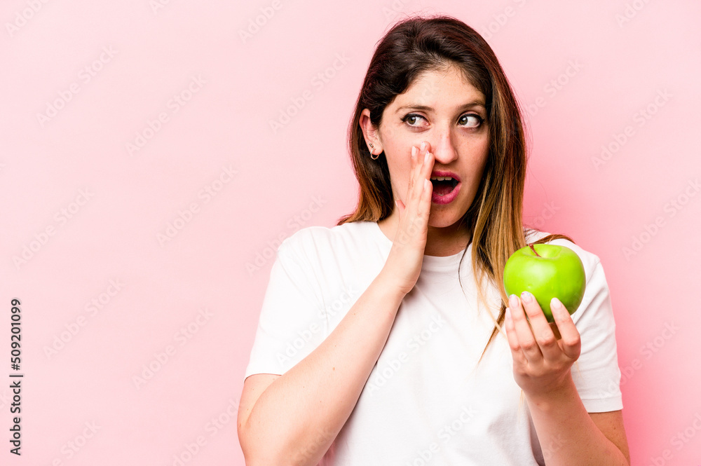 Young caucasian woman holding an apple isolated on pink background is saying a secret hot braking news and looking aside Young caucasian woman holding an apple isolated on pink background is saying a secret hot braking news and looking aside