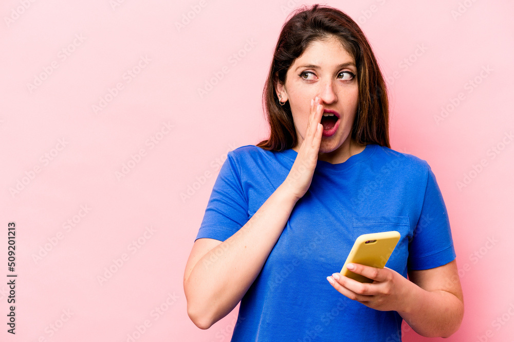 Young caucasian woman holding mobile phone isolated on pink background is saying a secret hot braking news and looking aside Young caucasian woman holding mobile phone isolated on pink background is saying a secret hot braking news and looking aside