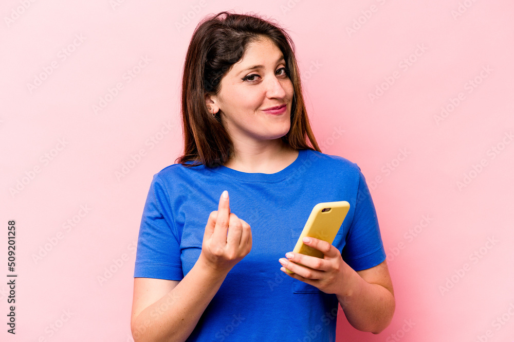 Young caucasian woman holding mobile phone isolated on pink background pointing with finger at you as if inviting come closer. Young caucasian woman holding mobile phone isolated on pink background pointing with finger at you as if inviting come closer.