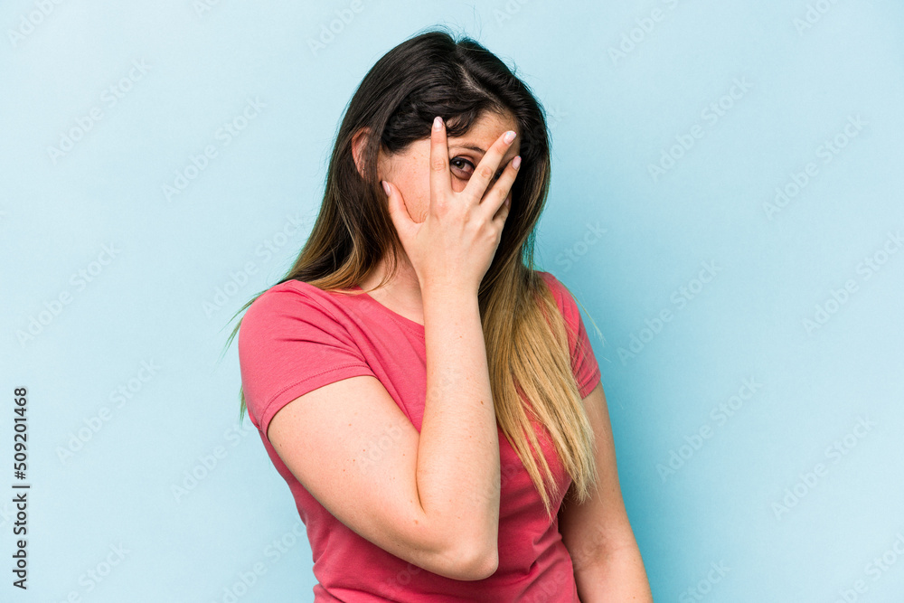 Young caucasian woman isolated on blue background blink at the camera through fingers, embarrassed covering face. Young caucasian woman isolated on blue background blink at the camera through fingers, embarrassed covering face.