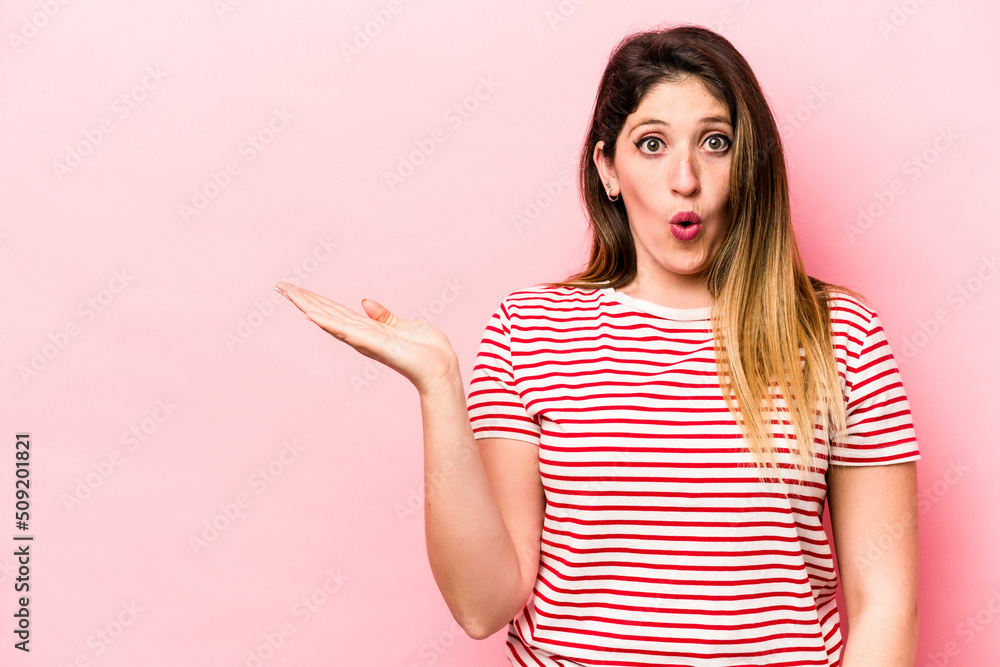 Young caucasian woman isolated on pink background impressed holding copy space on palm. Young caucasian woman isolated on pink background impressed holding copy space on palm.