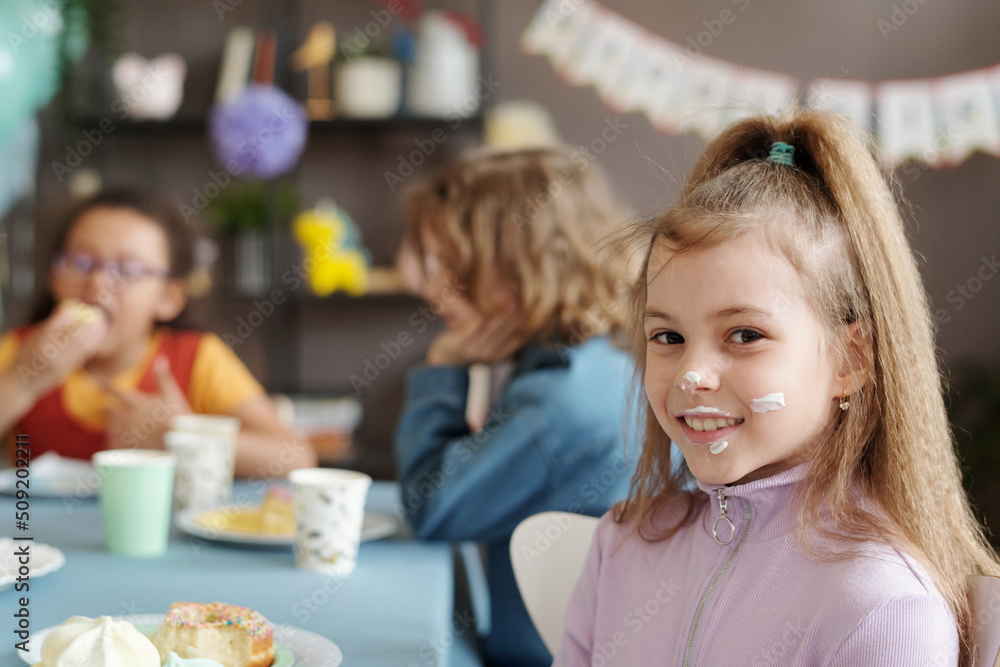 Portrait of little girl with cream on her face smiling at camera while sitting at table with her friends and eating dessert at party Portrait of little girl with cream on her face smiling at camera while sitting at table with her friends and eating dessert at party