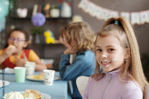 Portrait of little girl with cream on her face smiling at camera while sitting at table with her friends and eating dessert at party
