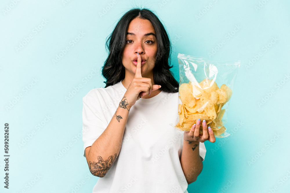 Young hispanic woman holding a bag of chips isolated on blue background keeping a secret or asking for silence. Young hispanic woman holding a bag of chips isolated on blue background keeping a secret or asking for silence.