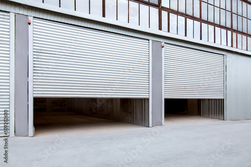 Opening gray iron shutter door of garage and industrial building warehouse exterior facade with grey concrete road, side view nobody.