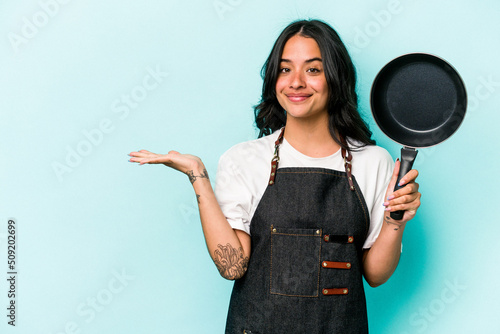 Fotótapéta Young hispanic cooker woman holding frying pan isolated on blue background showing a copy space on a palm and holding another hand on waist