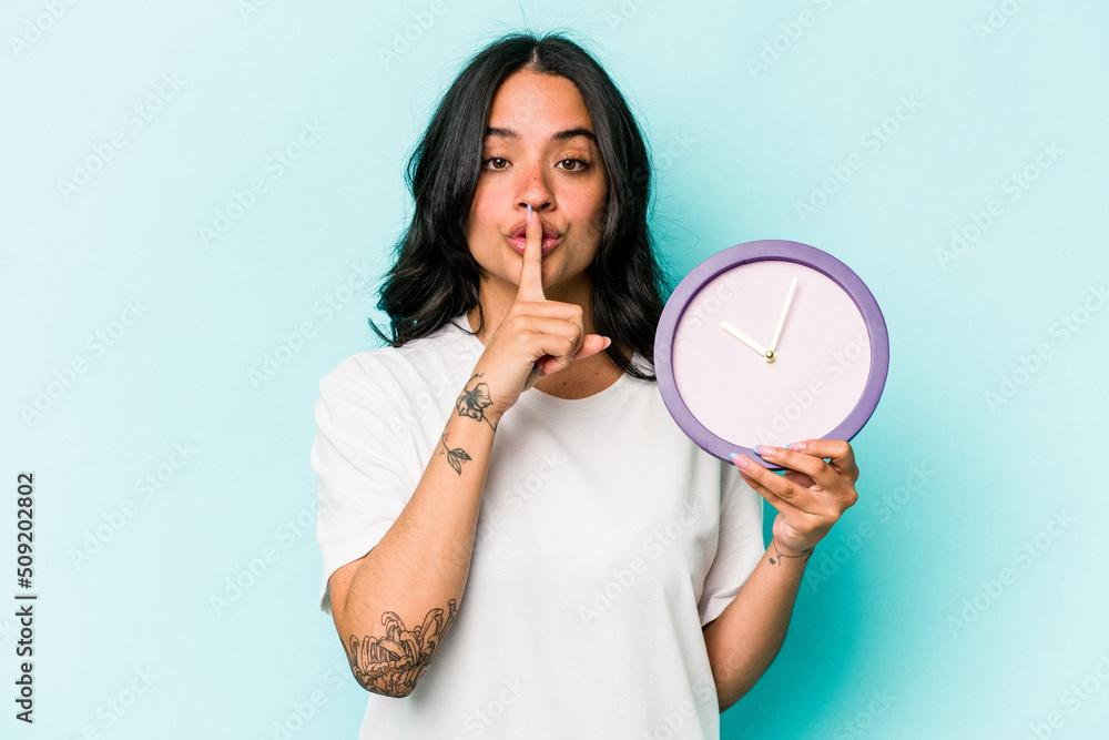Young hispanic woman holding a clock isolated on blue background keeping a secret or asking for silence. Young hispanic woman holding a clock isolated on blue background keeping a secret or asking for silence.