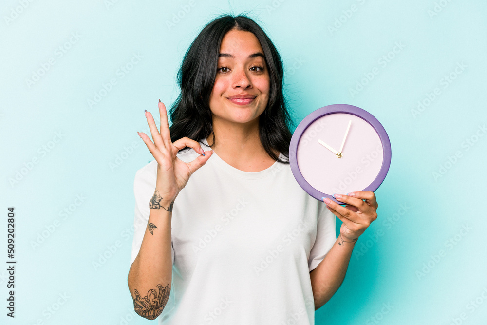 Young hispanic woman holding a clock isolated on blue background cheerful and confident showing ok gesture. Young hispanic woman holding a clock isolated on blue background cheerful and confident showing ok gesture.