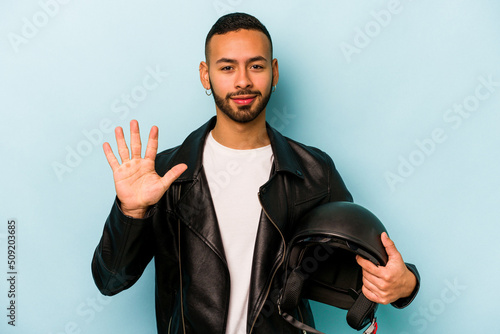 Young hispanic biker man isolated on blue background smiling cheerful showing number five with fingers.