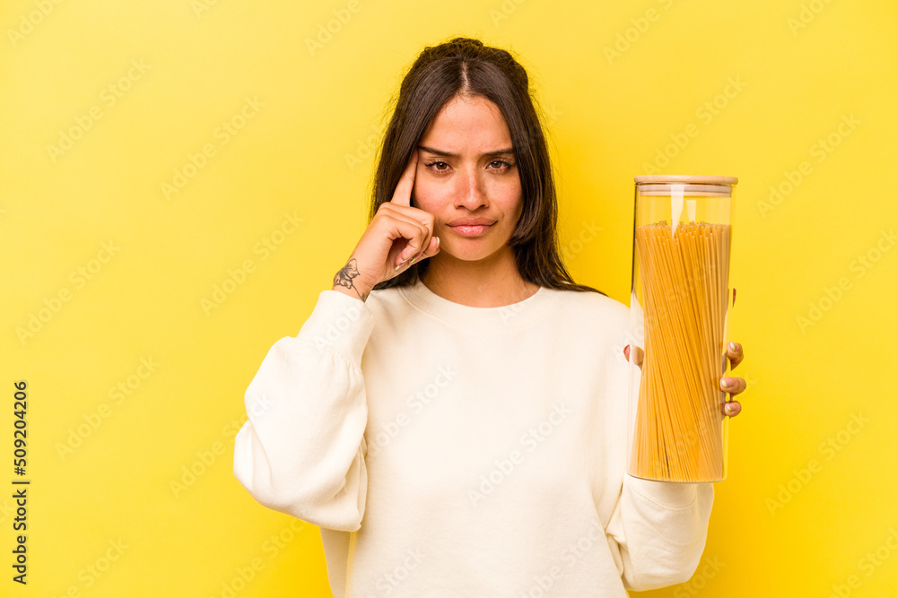 Young hispanic woman holding a spaghetti jar isolated on yellow background pointing temple with finger, thinking, focused on a task. Young hispanic woman holding a spaghetti jar isolated on yellow background pointing temple with finger, thinking, focused on a task.