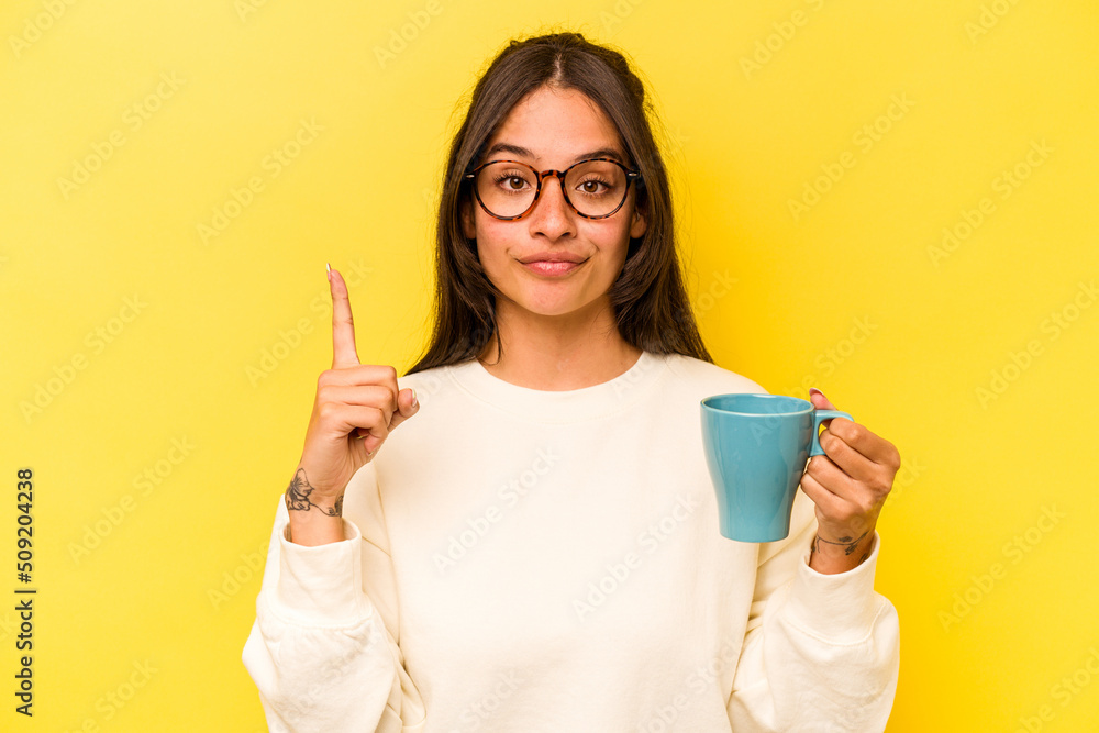 Young hispanic woman holding a mug isolated on yellow background showing number one with finger. Young hispanic woman holding a mug isolated on yellow background showing number one with finger.
