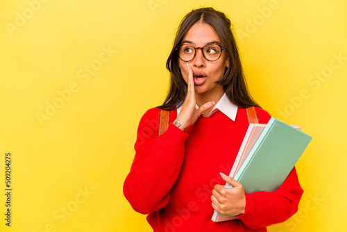 Young student hispanic woman isolated on yellow background is saying a secret hot braking news and looking aside
