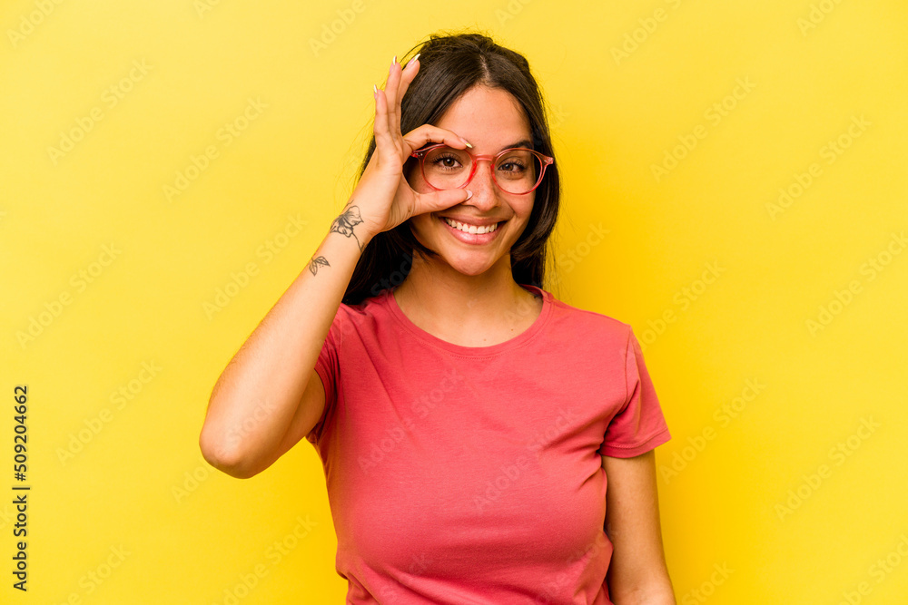 Young hispanic woman isolated on yellow background excited keeping ok gesture on eye. Young hispanic woman isolated on yellow background excited keeping ok gesture on eye.