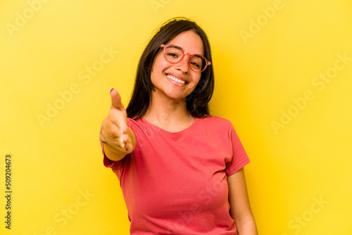 Young hispanic woman isolated on yellow background stretching hand at camera in greeting gesture.