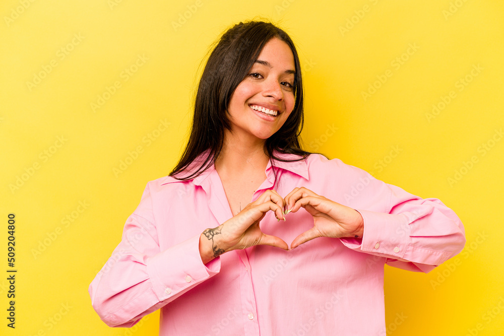 Young hispanic woman isolated on yellow background smiling and showing a heart shape with hands. Young hispanic woman isolated on yellow background smiling and showing a heart shape with hands.