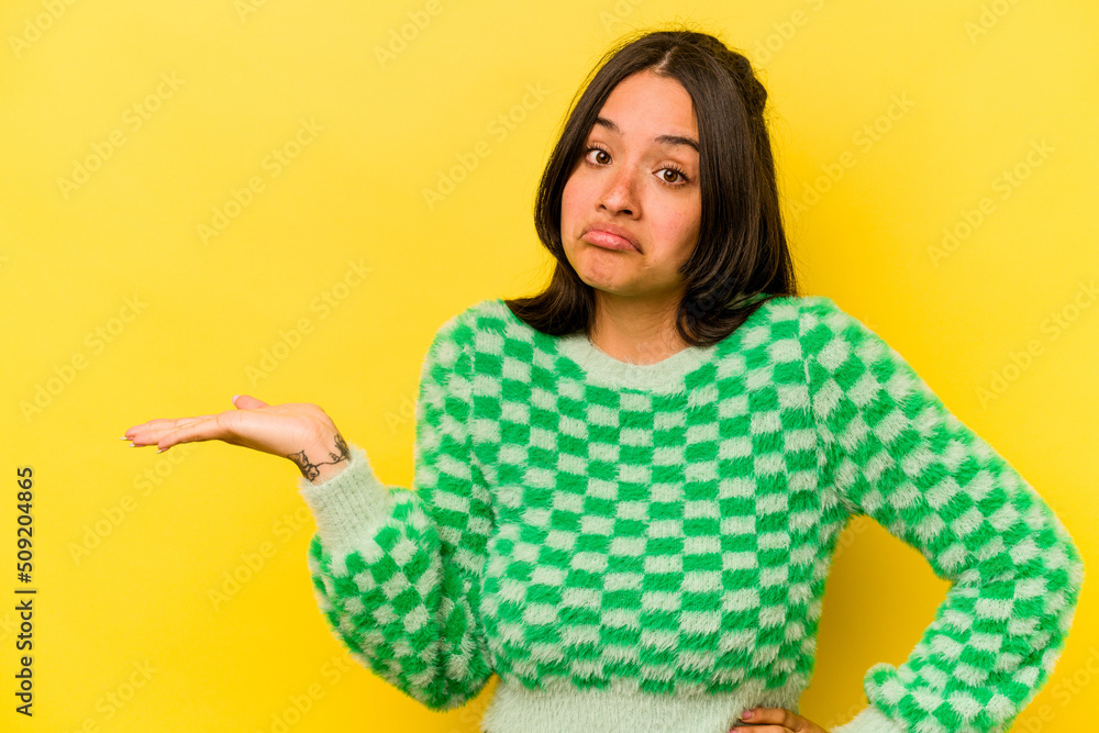 Young hispanic woman isolated on yellow background showing a copy space on a palm and holding another hand on waist. Young hispanic woman isolated on yellow background showing a copy space on a palm and holding another hand on waist.