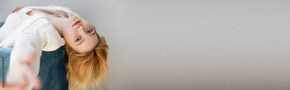Trendy young woman in jacket looking at camera on armchair isolated on grey, banner.