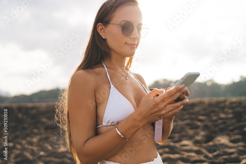 Millennial female tourist in sunglasses using cellular technology for chatting and messaging in social media networks during daytime at beach coastline, young woman writing content mobile publication