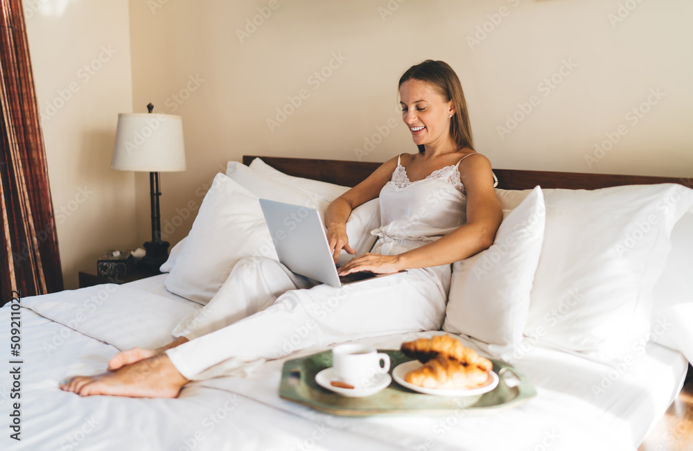Smiling female in silk nightgown using laptop application for booking hotel room service in online mode - enjoying weekend for yourself time, happy digital nomad chatting in netbook networks