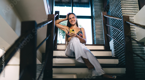 Portrait of cheerful Caucasian hipster girl sitting at stairs indoors and using mobile phone while smiling at camera, cheerful millennial woman holding mobile phone enjoying youth lifestyle