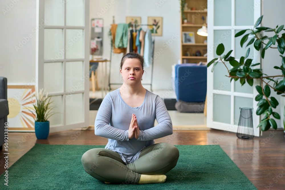 Young woman with disability sitting in pose of lotus on the floor of spacious living room and looking at camera while practicing yoga Young woman with disability sitting in pose of lotus on the floor of spacious living room and looking at camera while practicing yoga