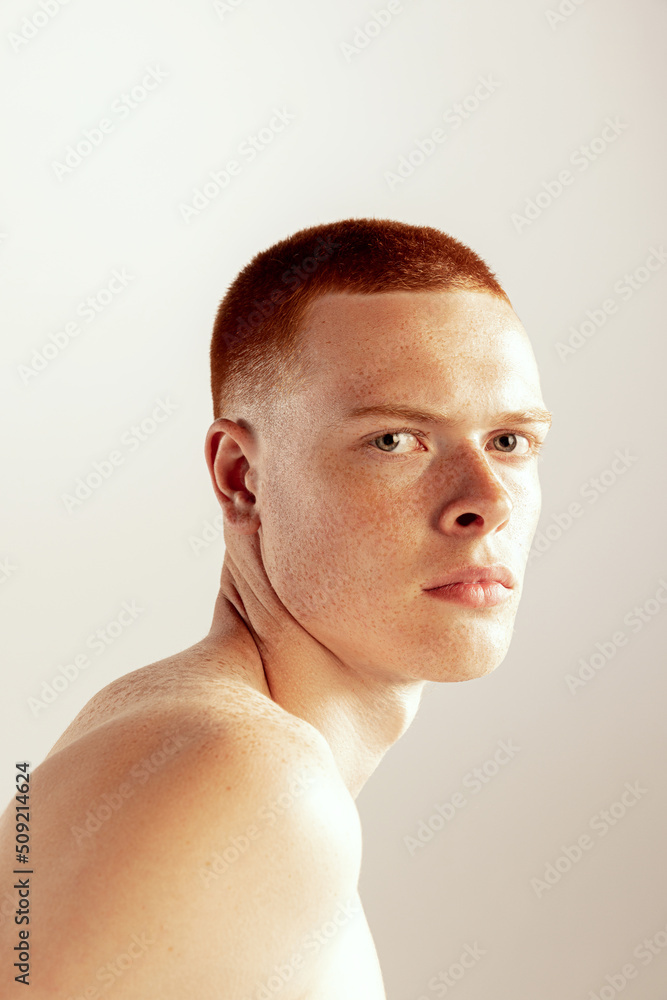 Portrait of young red-haired man with freckled face posing, seriously looking at camera isolated over grey studio background. Side view Portrait of young red-haired man with freckled face posing, seriously looking at camera isolated over grey studio background. Side view