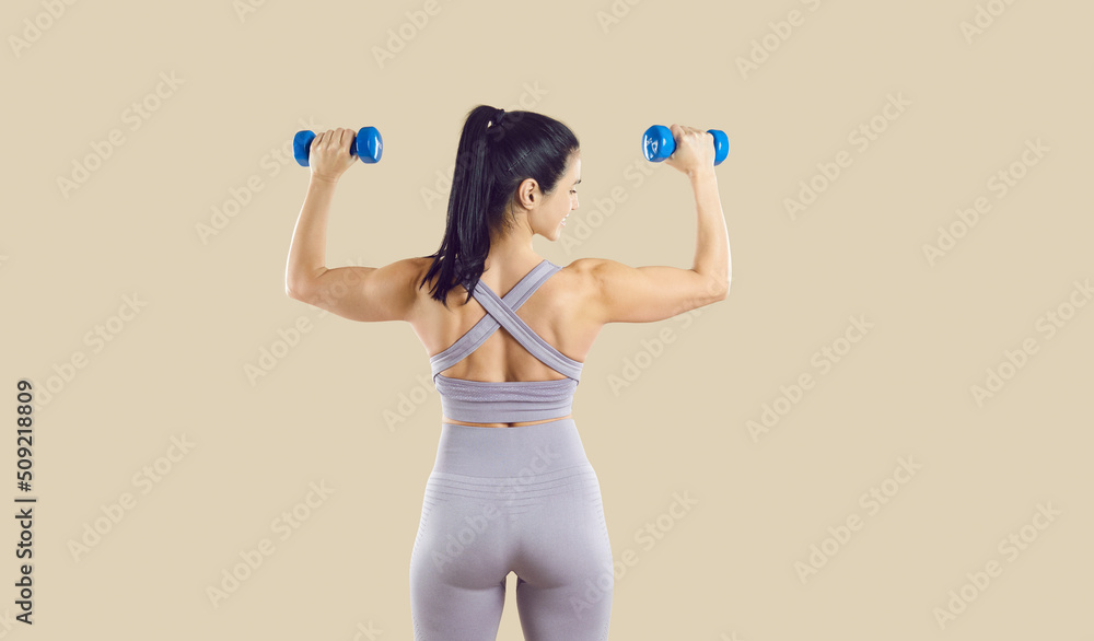 Happy sporty woman lifts two small dumbbells while standing with her back to camera on beige background. Rear view of young brunette woman with beautiful figure dressed in stylish gray sportswear. Happy sporty woman lifts two small dumbbells while standing with her back to camera on beige background. Rear view of young brunette woman with beautiful figure dressed in stylish gray sportswear.