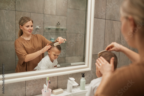 Portrait of caring Caucasian mother combing hair or mixedrace baby boy by mirror in bathroom, copy space photo