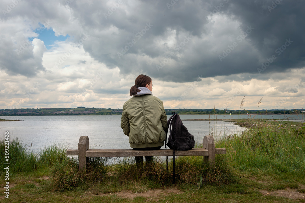 Fototapeta premium Rear view of a woman sitting on a bench alone looking at the view across the water, stormy sky.