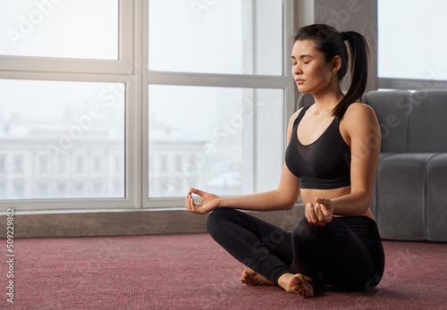 Side view of young, brunette girl in black sports top sitting in easy pose, meditating in light room. Barefoot girl practicing sukhasana with closed eyes, alone on carpet. Concept of meditation yoga.