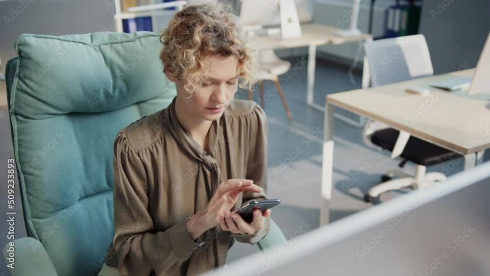 Shot of serious female Chief Editor using smartphone, calling to colleagues and discussing work. Smart Caucasian woman with curly hair sitting at workplace in cozy office. Daytime Shot of serious female Chief Editor using smartphone, calling to colleagues and discussing work. Smart Caucasian woman with curly hair sitting at workplace in cozy office. Daytime