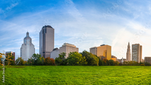 Panoramic Downtown Hartford Cityscape over the green park with the blue sky and white clouds at sunrise. Beautiful tranquil morning landscape of modern downtown buildings in natural settings.