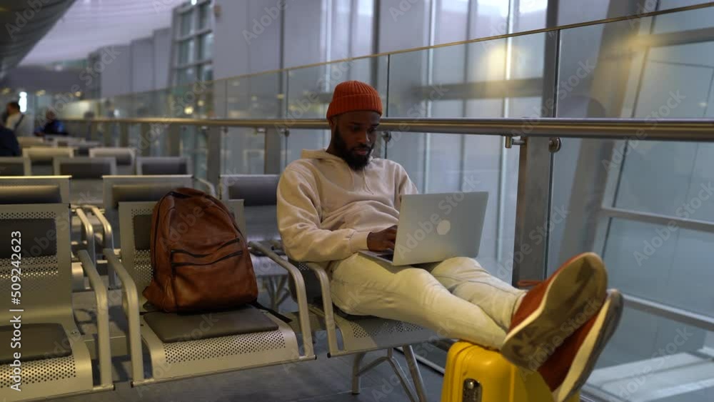 Focused young African American man sitting in hall of airport terminal working on laptop. Concentrated freelancer black male online working on computer and waiting landing on board. Business travel. Focused young African American man sitting in hall of airport terminal working on laptop. Concentrated freelancer black male online working on computer and waiting landing on board. Business travel.