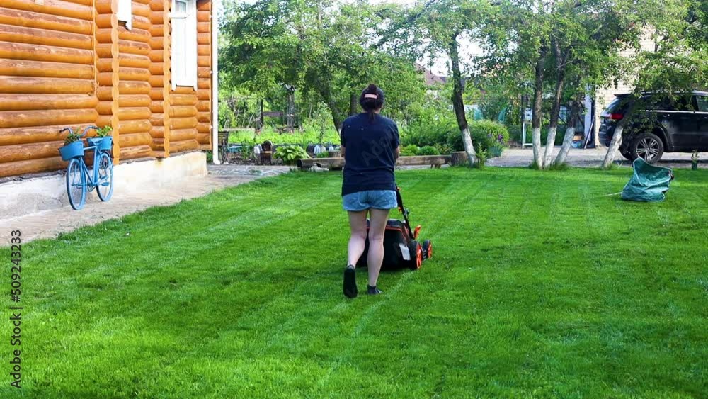 Woman mowing green lawn in backyard near house and car moving away area with lawn mower on summer sunny day back view, slow motion. Landlady cleaning up territory. Care and maintenance of home garden.