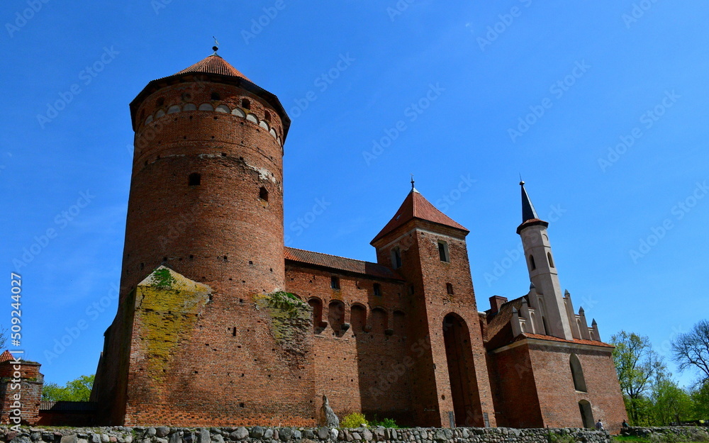 A close up on an old medieval castle made out of red brick, with a  tall tower, angled roof and some addition in the shape of a spire seen on a sunny summer day on a Polish countryside