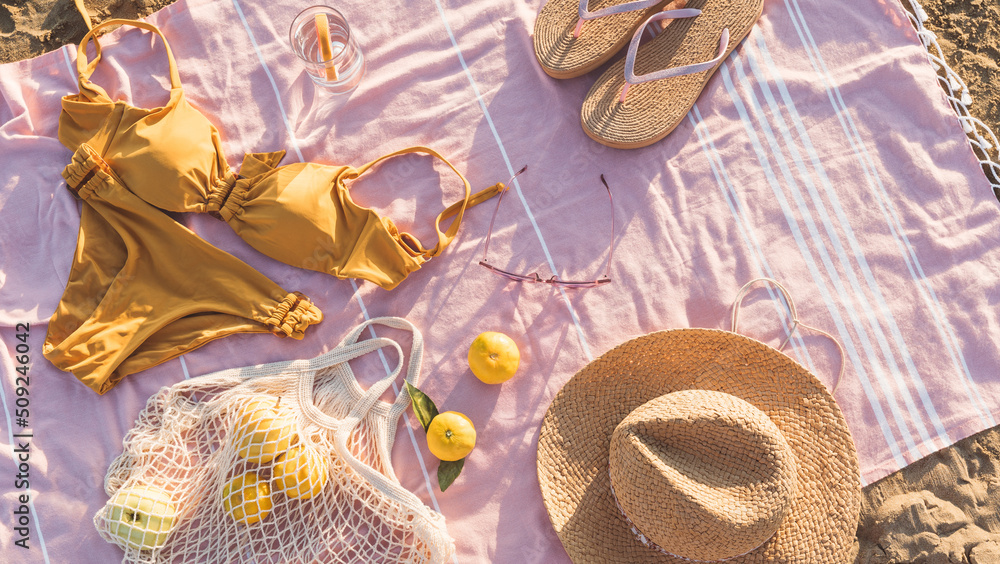 Summer beach accessories flat lay on sand background. Holiday travel, tropical concept. Straw hat, bikini, sunglasses, slippers and fruits in eco friendly mesh shopping bag. Sun shadow and sunlight
