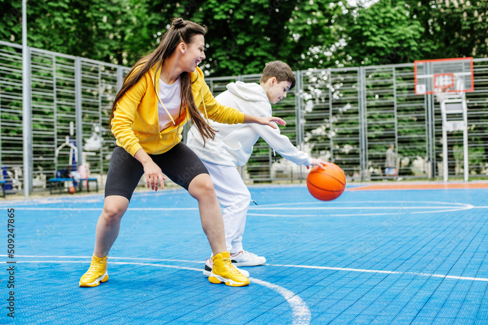 Obraz premium Girl and her younger brother, teenager, play basketball on modern basketball court under open sky