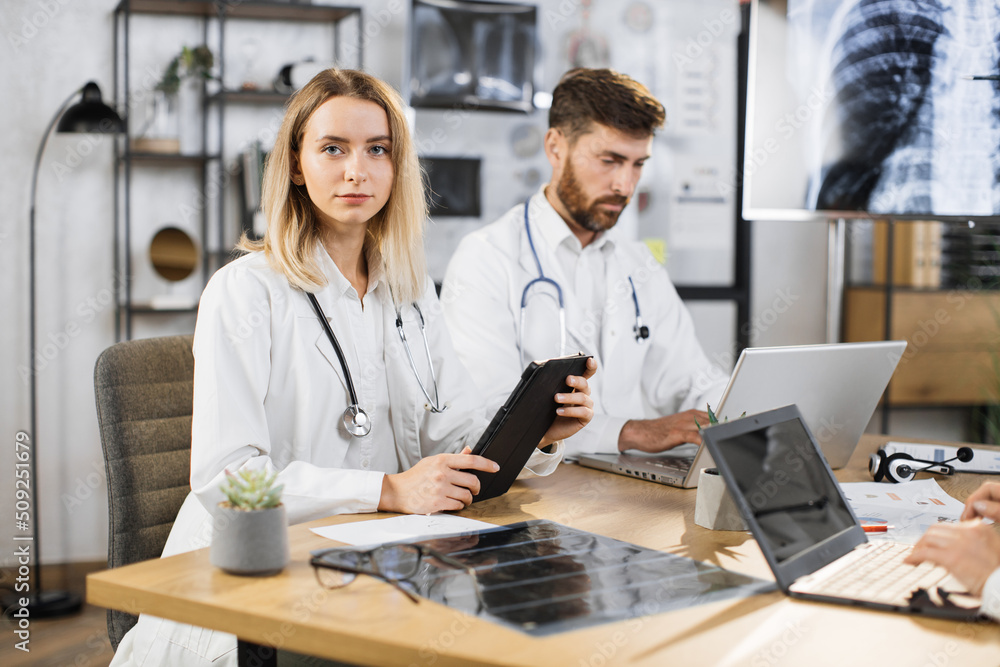 Portrait of caucasian female doctor sitting at table during medical staff meeting with digital tablet in hands. Bearded concentrated male physician working on laptop on background. Portrait of caucasian female doctor sitting at table during medical staff meeting with digital tablet in hands. Bearded concentrated male physician working on laptop on background.