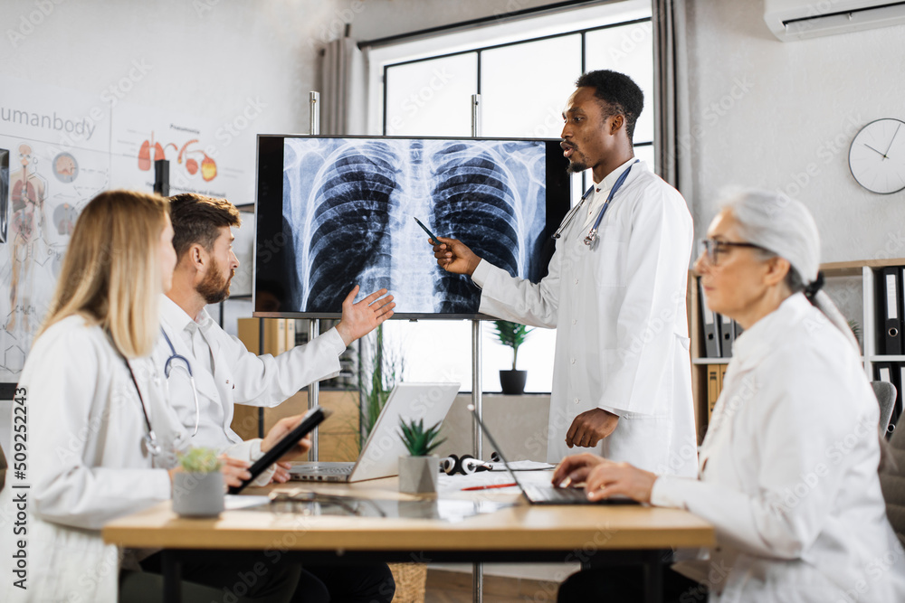 Four multicultural doctors looking at digital screen with x-ray of lungs during medical staff meeting. African american physician arguing with caucasian bearded colleague about patient diagnosis. Four multicultural doctors looking at digital screen with x-ray of lungs during medical staff meeting. African american physician arguing with caucasian bearded colleague about patient diagnosis.