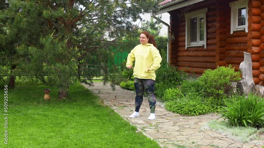 Red-haired fat and overweight woman in yellow sweatshirt doing breathing exercises, fitness, squatting in the green backyard outdoors. Countryside, wooden door on background. Losing weight, plus size Red-haired fat and overweight woman in yellow sweatshirt doing breathing exercises, fitness, squatting in the green backyard outdoors. Countryside, wooden door on background. Losing weight, plus size