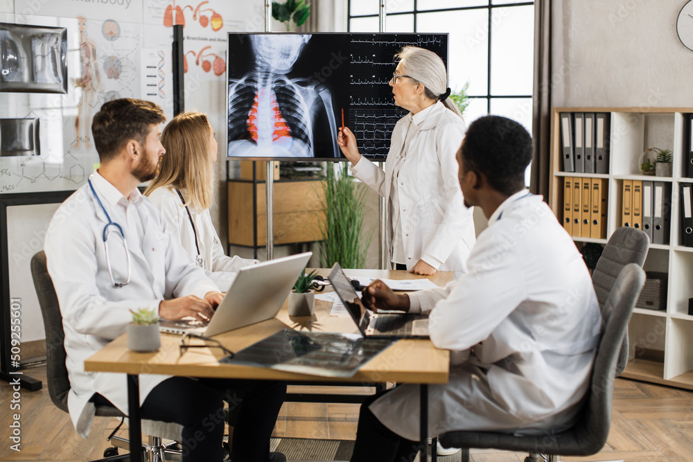 Obraz premium Aged medical worker discussing with multiracial colleagues x ray scan and heartbeat results shown on big digital screen. Team of doctors having briefing at boardroom about severe patient.