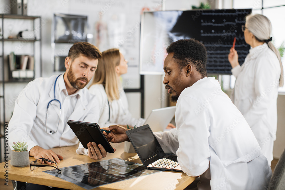 Fototapeta premium Two male doctors sitting at desk and using laptop with tablet while female specialists examining x ray scan on digital monitor. Group of international medical workers having meeting at boardroom .
