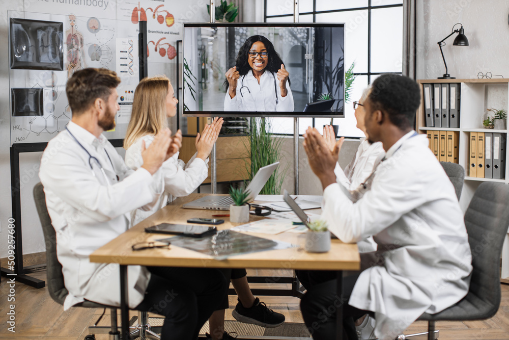 Team of multicultural doctors in white lab coats clapping hands during online video conference after successful speech of excited female scientist. Focus on african american woman on monitor screen. Team of multicultural doctors in white lab coats clapping hands during online video conference after successful speech of excited female scientist. Focus on african american woman on monitor screen.