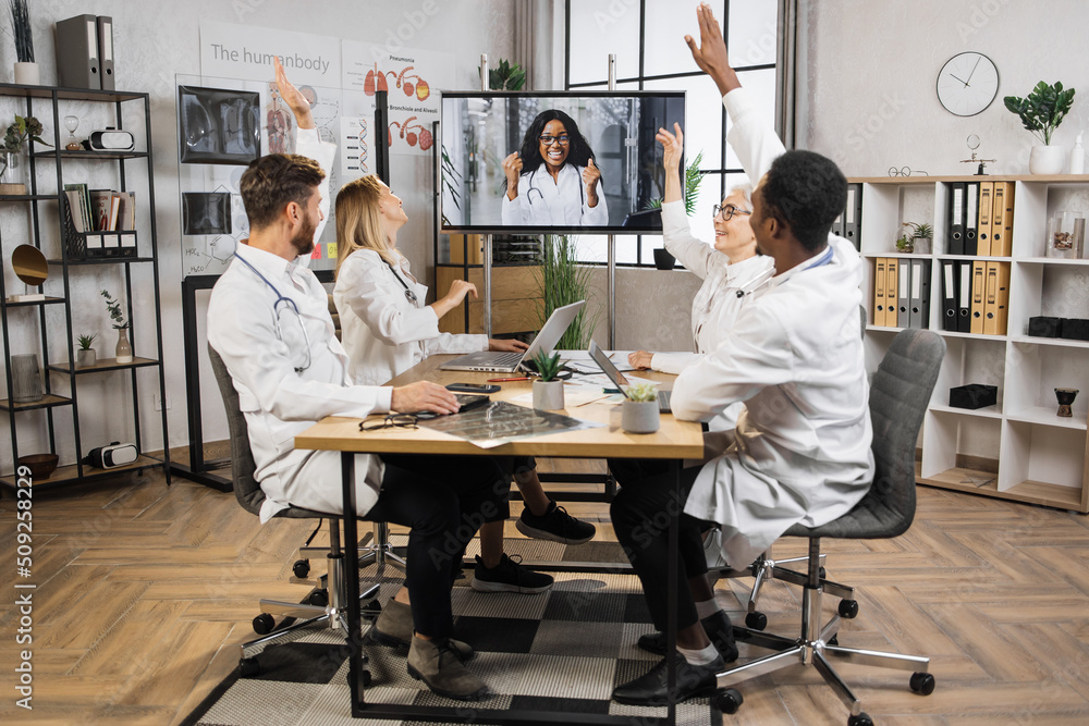 Group of motivated international scientists sitting at table and raising hands in hospital conference room. Excited multiethnic workers unanimously voting for suggested idea during online video call. Group of motivated international scientists sitting at table and raising hands in hospital conference room. Excited multiethnic workers unanimously voting for suggested idea during online video call.
