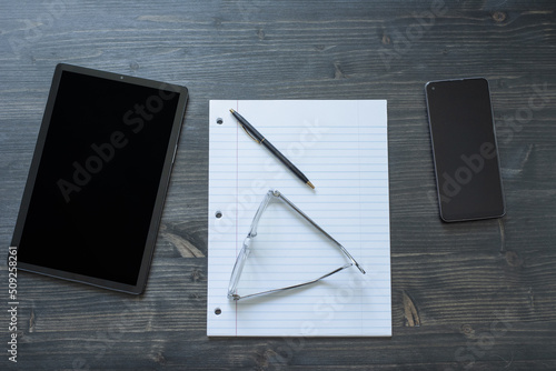 Desk top from top view looking at tablet, paper, pen, phone, and pair of glasses.